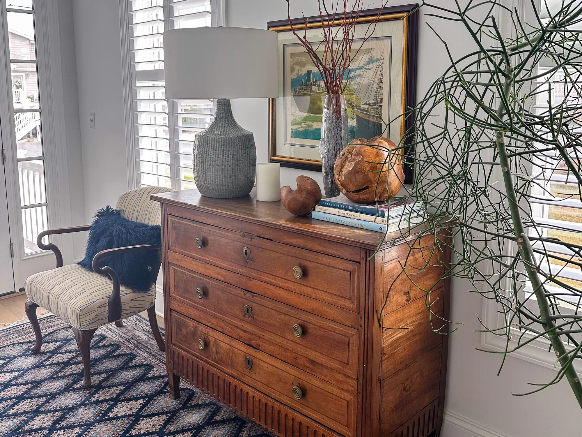 A wood dresser with a gray lamp, books, and a framed print beside a striped chair
