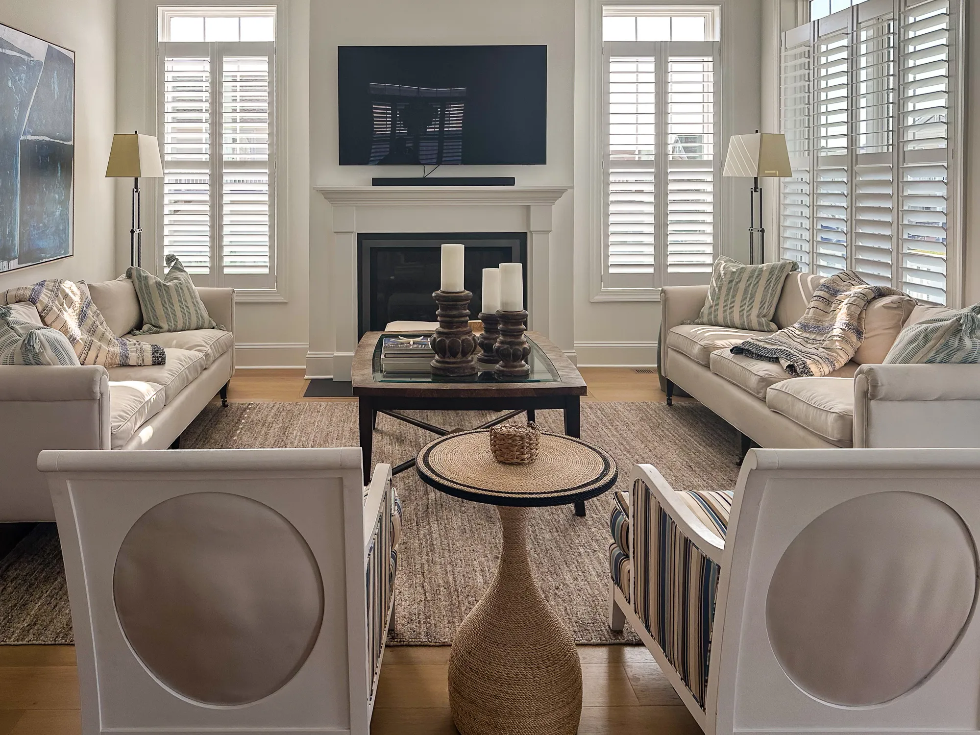 A living room with white sofas, a glass coffee table, and shuttered windows